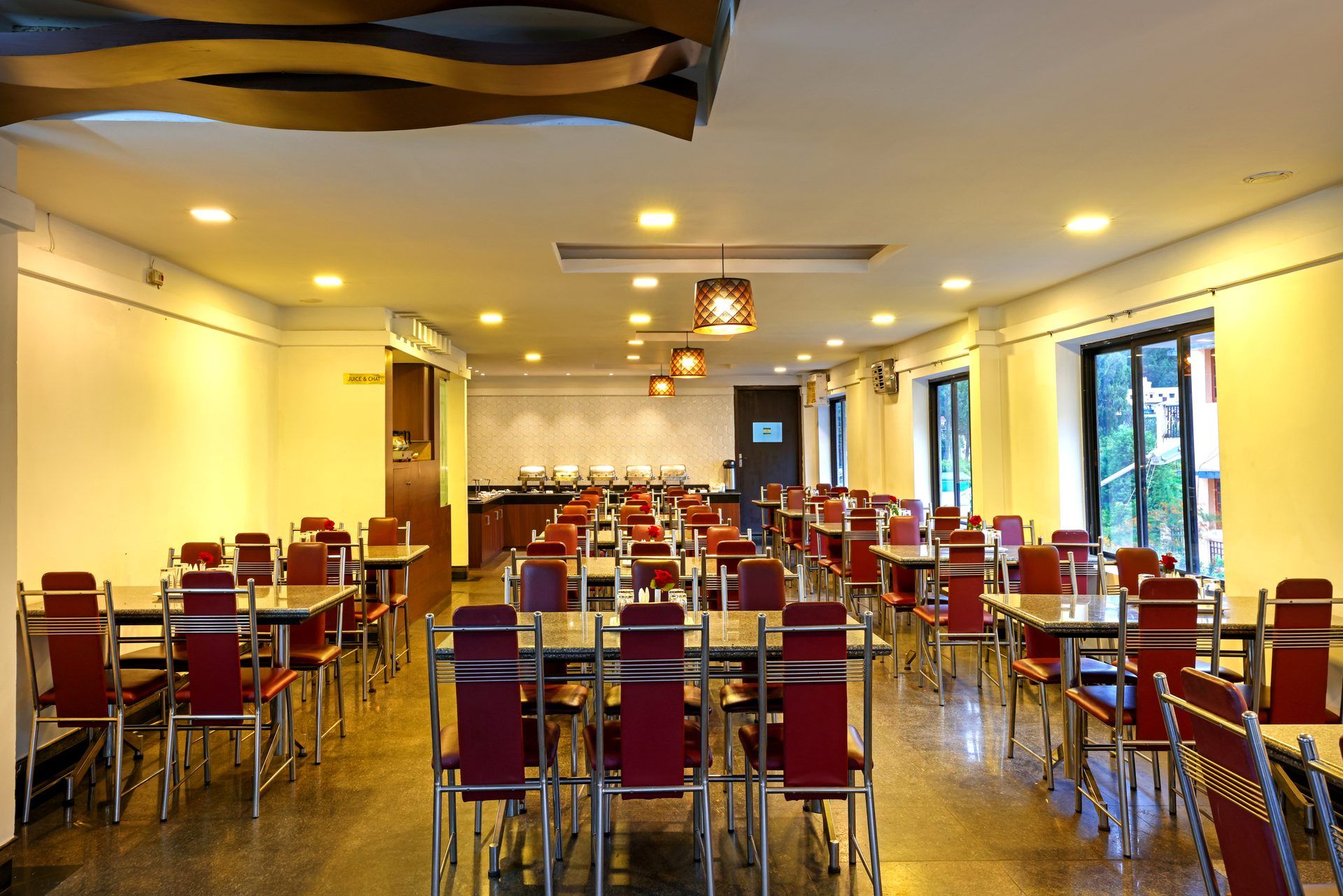 Uma sala de jantar ampla, com várias mesas e cadeiras, um buffet e grandes janelas que oferecem luz natural.