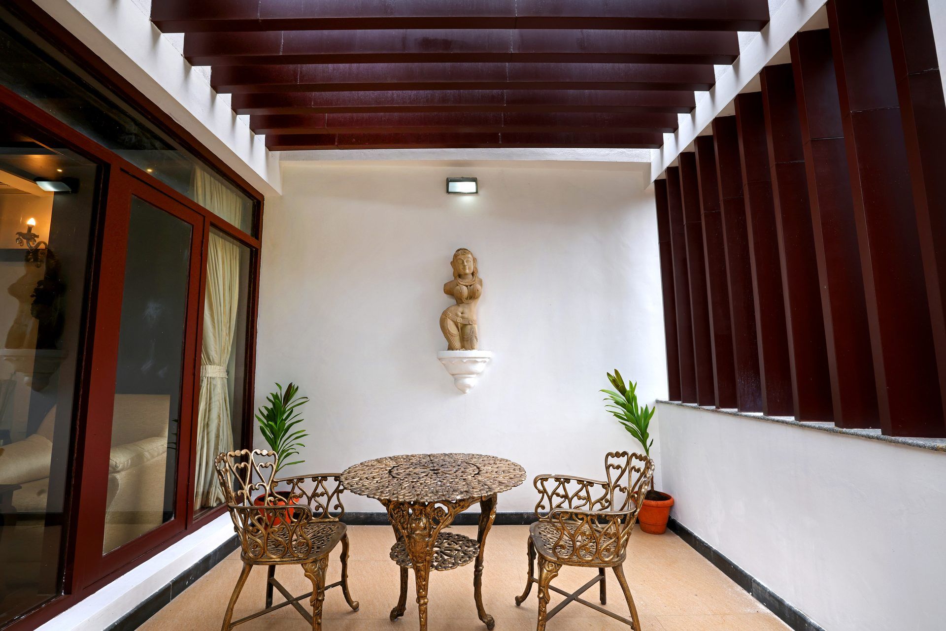 Uma área de varanda coberta com uma mesa redonda e duas cadeiras, uma escultura na parede e plantas em vasos.