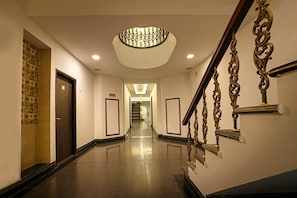 Un pasillo de hotel con piso pulido, una imponente escalera con barandillas de metal ornamentadas y una lámpara decorativa en el techo.