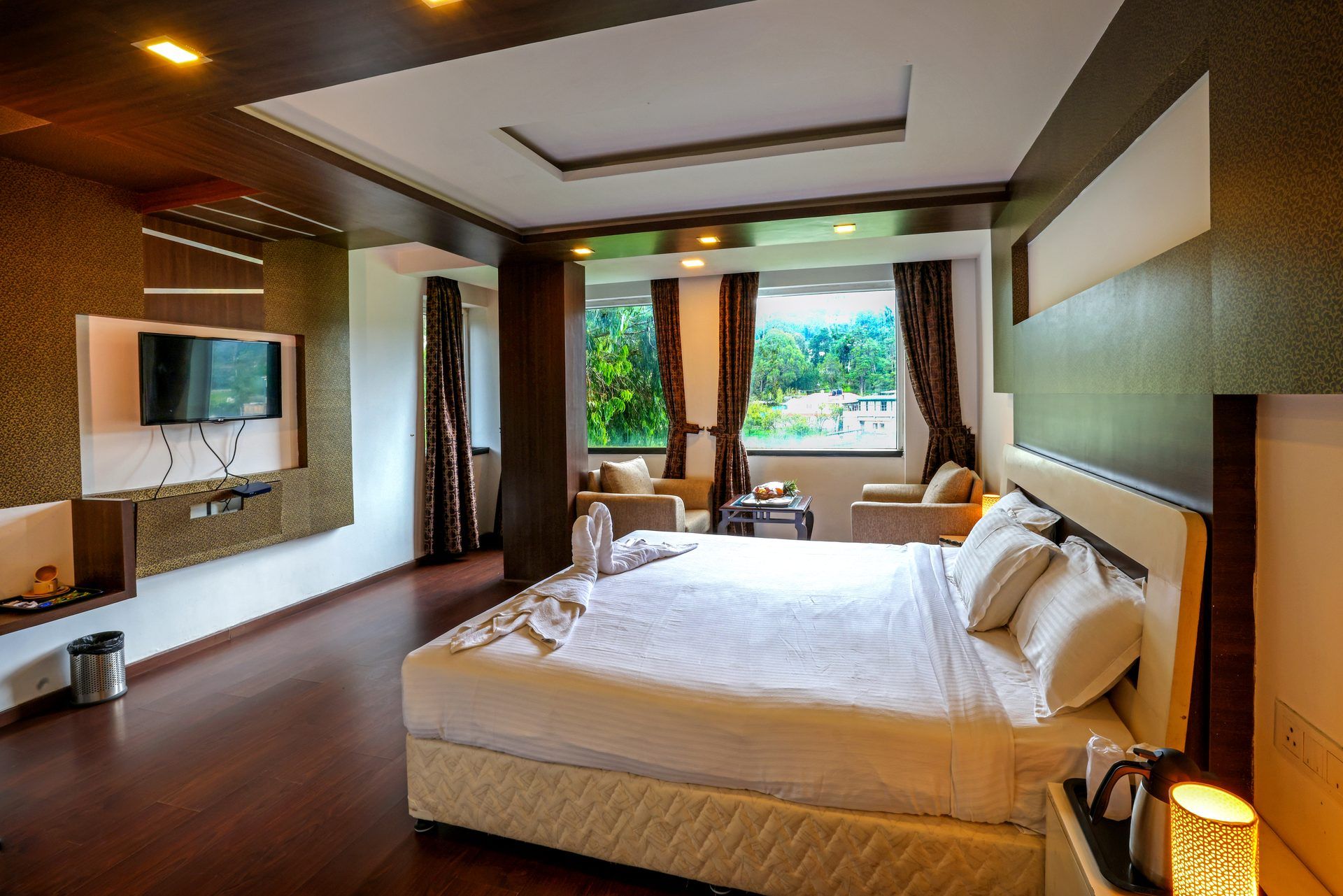 Habitación de hotel moderna con una cama grande, un televisor de pantalla plana en la pared, una mesita con una bandeja de fruta y vistas a zonas verdes.