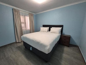 Un dormitorio con cama, mesita de noche, ventana con cortinas y lámpara.