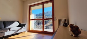 A room with a large sliding glass door leading to a balcony, a grey sofa, and a small wooden table with a stuffed animal. - Tommy's Cottage in Aprica - Mountain views and ski lifts just a short walk away (Corteno Golgi)