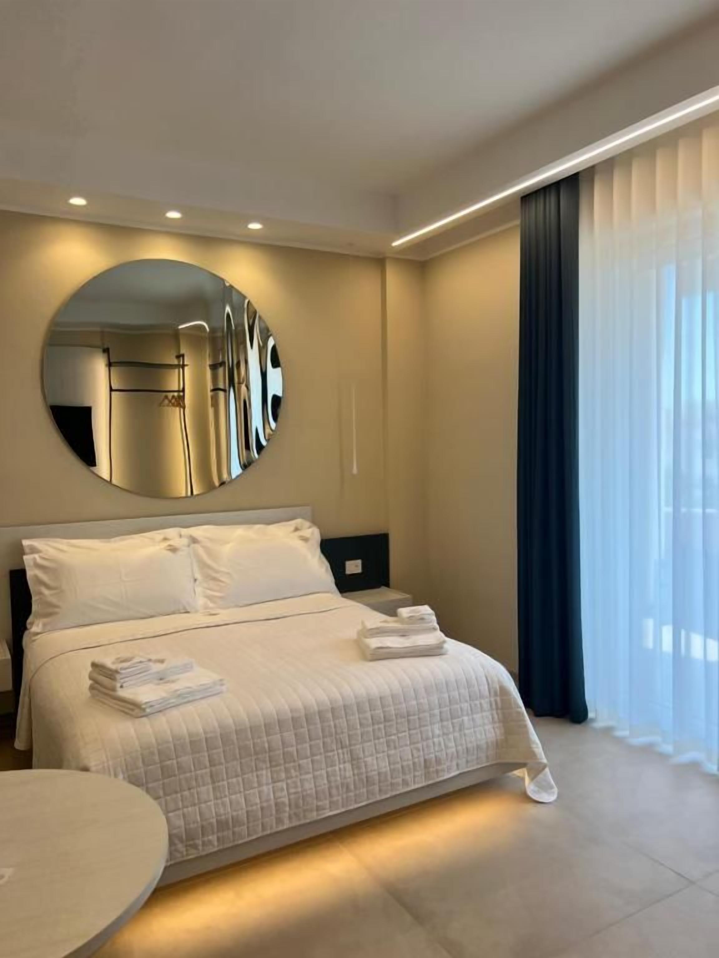 Ein modernes Hotelzimmer mit einem großen Bett, einem runden Spiegel, einem Nachttisch und Blick ins Freie durch transparente Vorhänge.