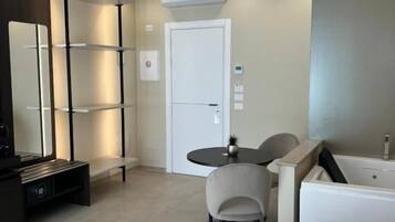 A modern hotel room with a bed, a small table, a chair, a desk, a shelf, a mirror, a door, and a bathtub.