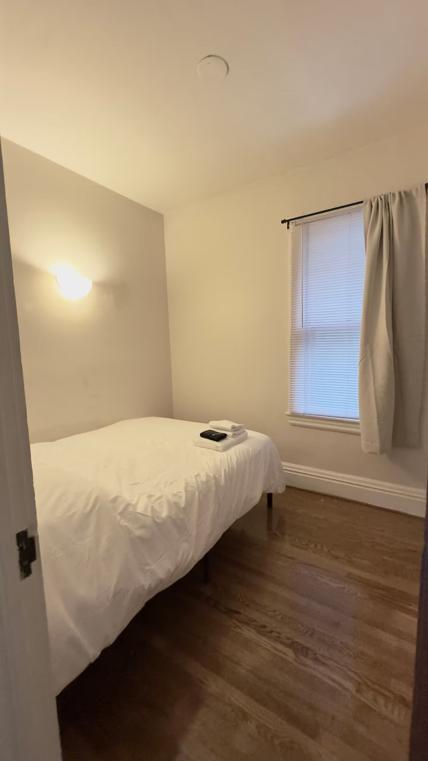 Een slaapkamer met een enkel bed, een nachtkastje, een raam met zonwering en een lamp aan de muur.