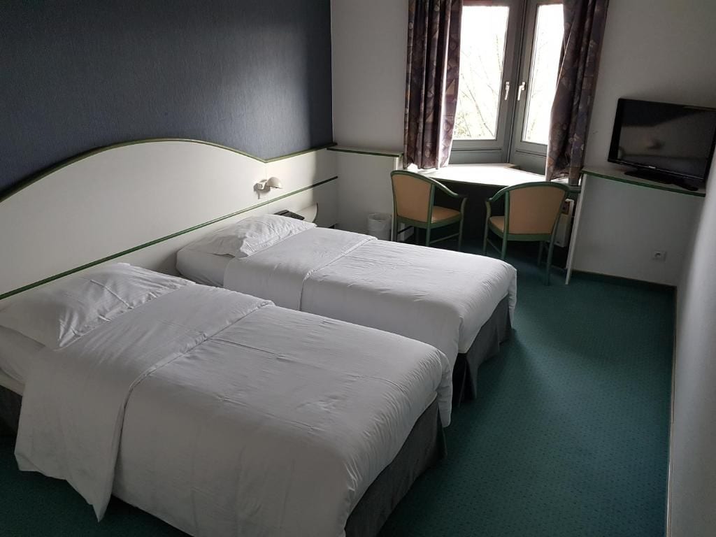 A hotel room with two beds, a desk with chairs, a television, and a window with curtains.