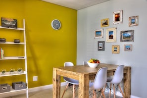 A dining area with a wooden table, white chairs, and a bowl of fruit. A white shelf with decorative items is against a yellow wall. - Serene 1-bedroom apartment in Cape Town perfect for relaxing stays (Cape Town)