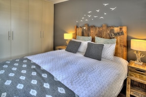 A bed with a quilted bedspread, two pillows, a nightstand with a lamp, and a headboard with a wooden design and a heart-shaped decoration. - Serene 1-bedroom apartment in Cape Town perfect for relaxing stays (Cape Town)
