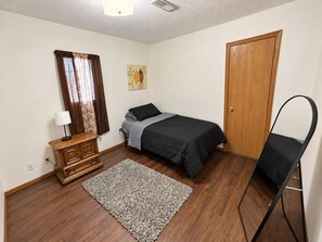 Un dormitorio con una cama, un aparador de madera, un escritorio con una lámpara, una puerta y una silla.