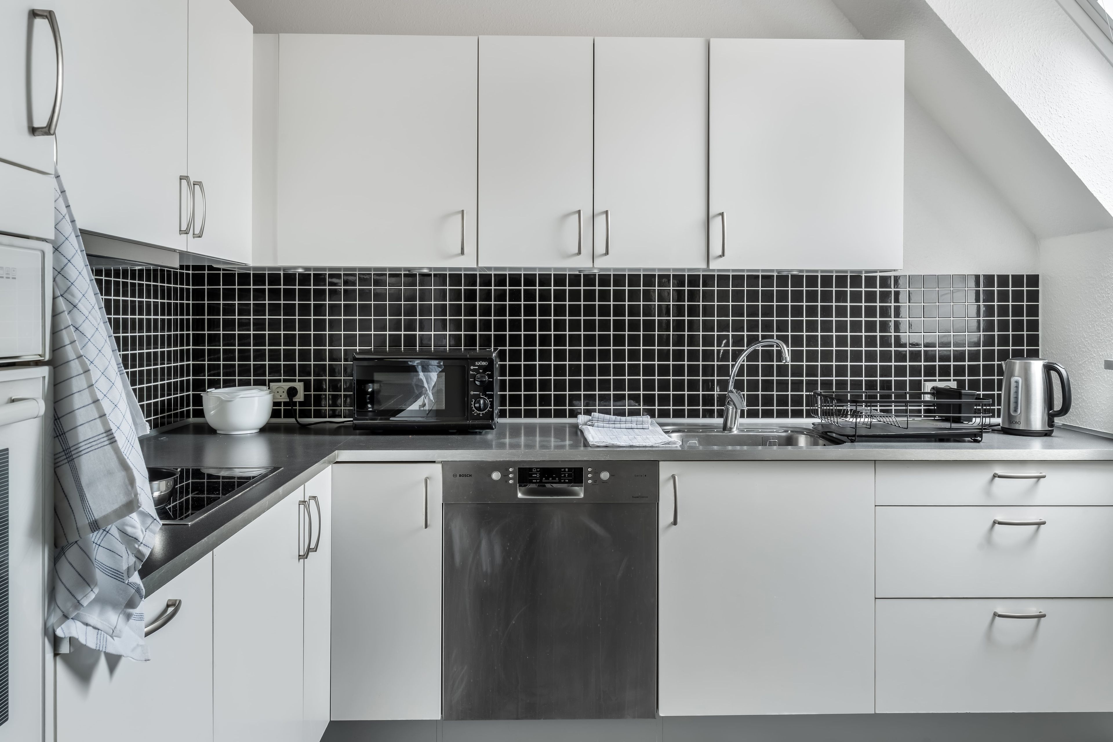 Una cocina moderna con armarios blancos, salpicadero de azulejos negros, electrodomésticos de acero inoxidable y lavavajillas.