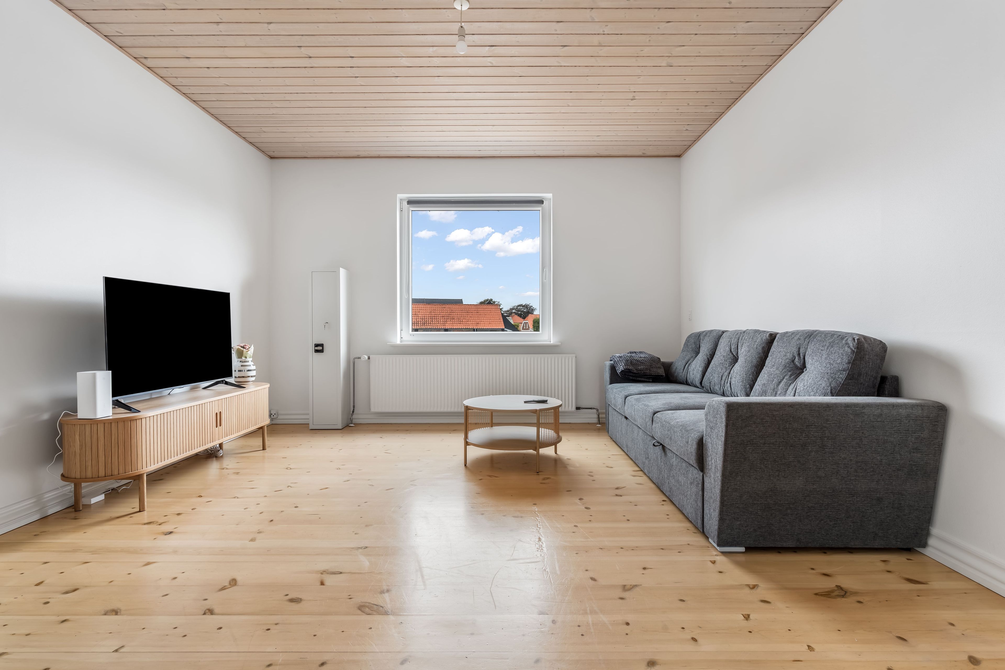 Uma sala de estar moderna com televisão de tela plana, um sofá cinza, uma mesa redonda pequena e uma janela com vista para um edifício de telhado vermelho.