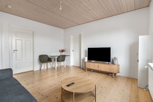 A modern living room with a wooden floor, a round coffee table, a grey sofa, a TV on a wooden cabinet, and a small table with chairs.
