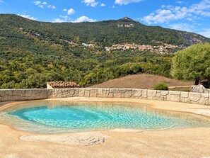 Ein Schwimmbecken mit klarem Wasser, umgeben von einer Steinmauer, mit Blick auf einen Hügel und Gebäude.