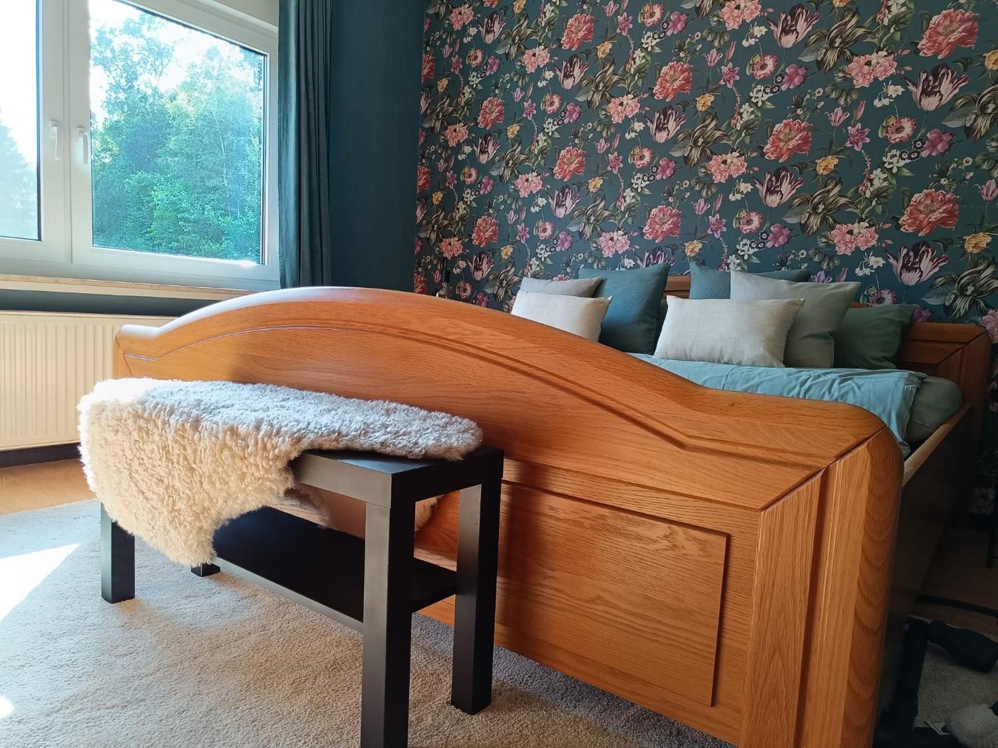 Ein Schlafzimmer mit einem hölzernen Kopfteil, einer flauschigen weißen Decke auf einem Sessel, einem Bett mit vielen Kissen und floralem Wandpapier.