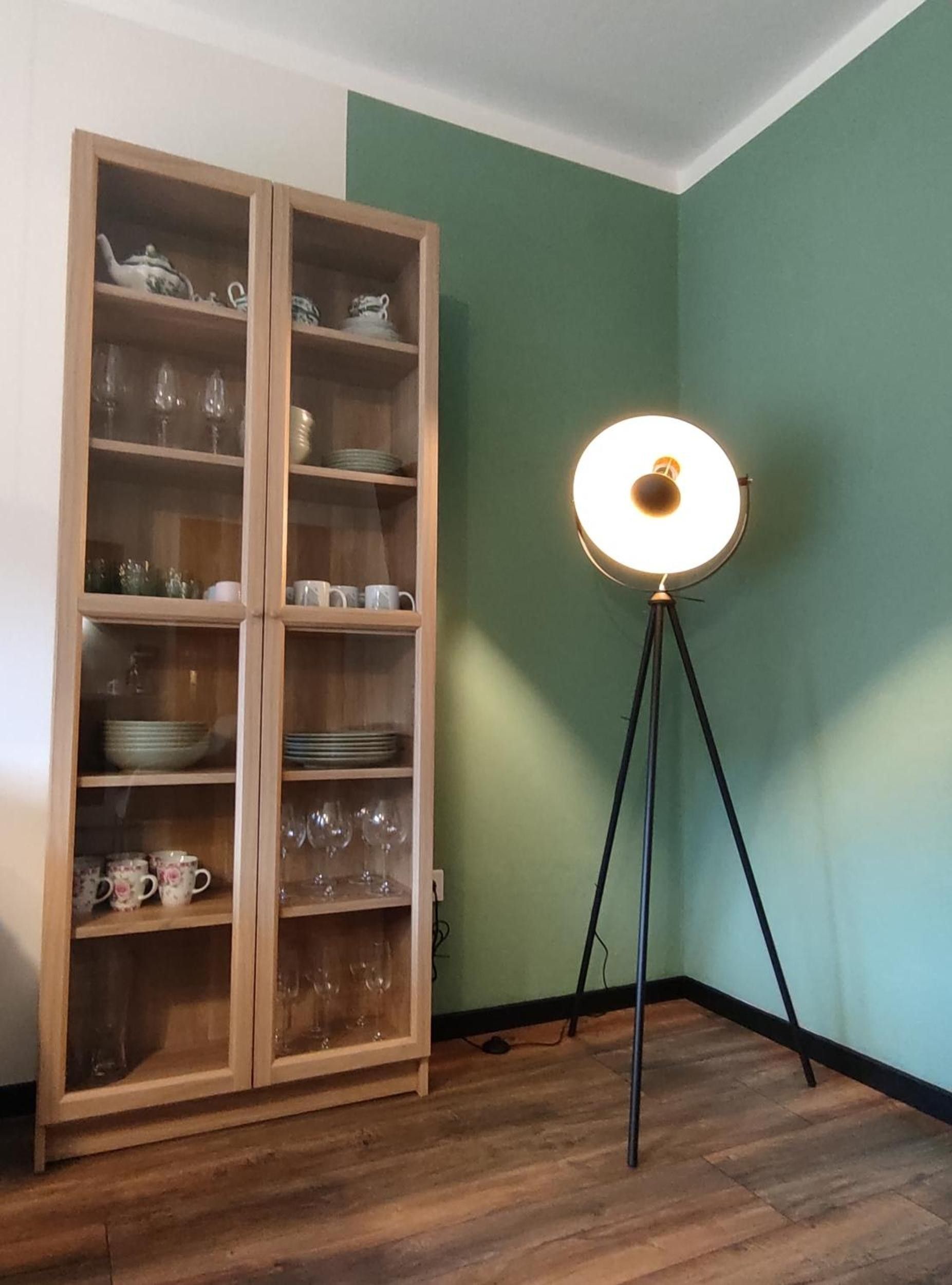 Ein Zimmer mit einem Holzschrank, der Glaswaren und Geschirr beherbergt, einer Stehlampe und einer grünen Wand.