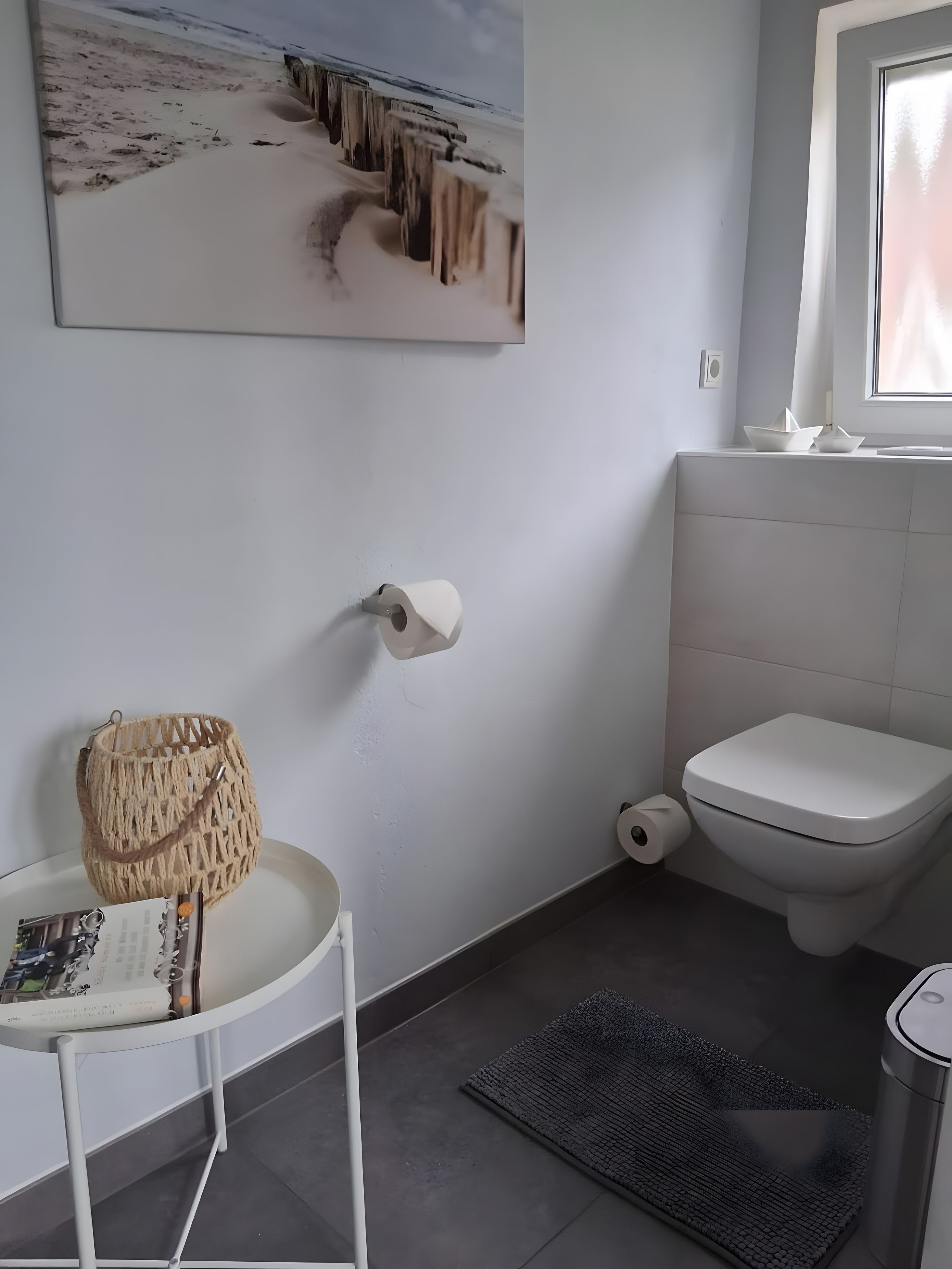 A bathroom with a toilet, a roll of toilet paper, a small table with a magazine, a decorative basket, and a rug on the floor.