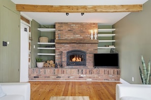 A living room with a brick fireplace, wooden beams, a flat-screen TV, and a white sofa. - The Halliday Creek Cabin (200 Acres of Privacy) (Greater Madawaska)