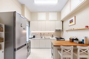 A modern kitchen with white cabinetry, a stainless steel refrigerator, a wooden dining table with white chairs, and a small island with a sink. - Seafront Apartment | Panoramic Sea Views | Thessaloniki Center (Thessaloniki)