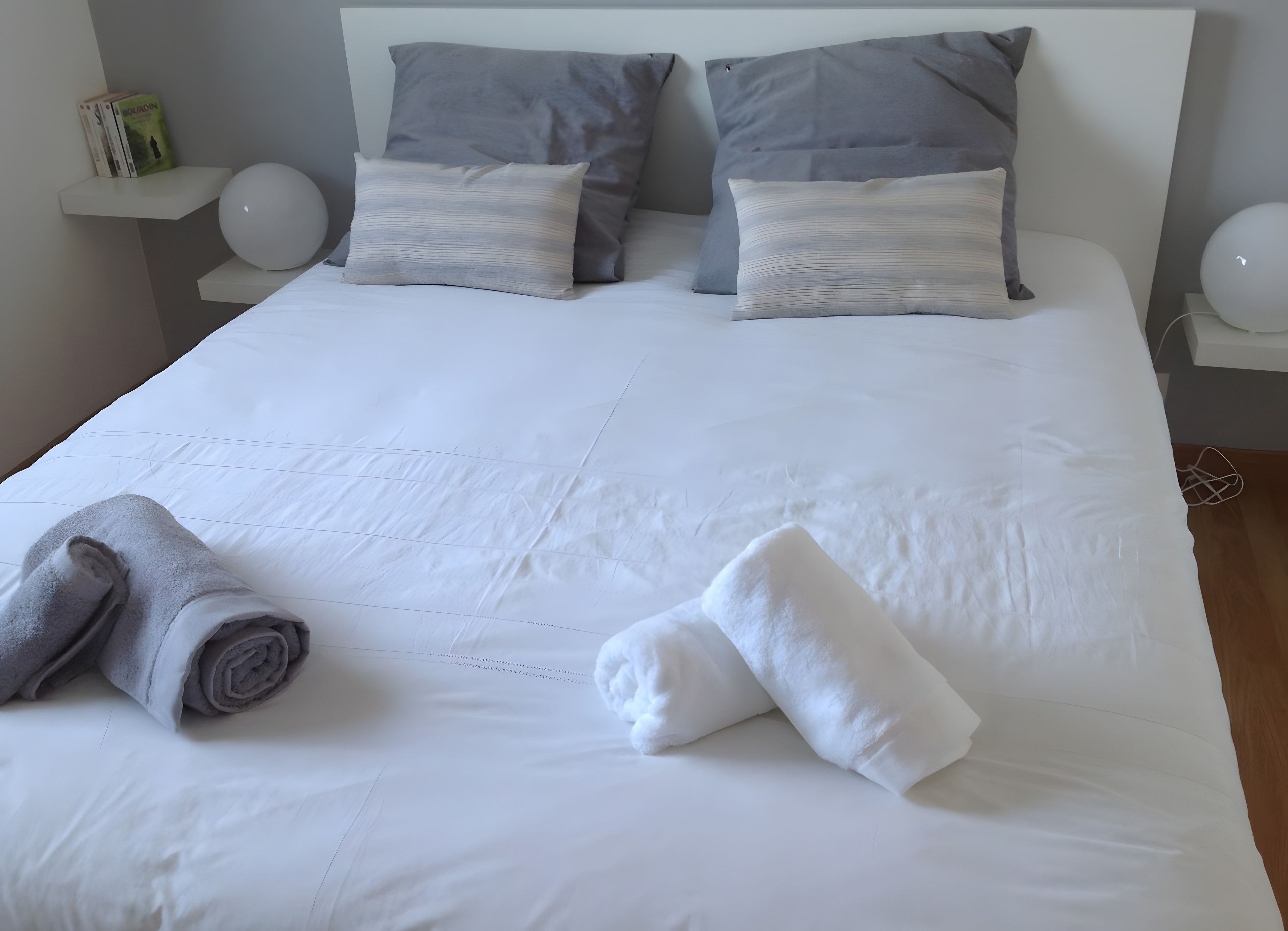 A neatly made bed with pillows, a rolled towel, and a bedside table with a book and a lamp.