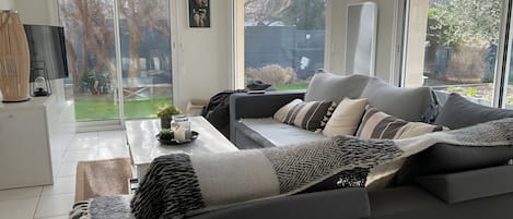 Een moderne woonkamer met een grijze bank, een bijzettafeltje met decoratieve objecten, en een groot raam met uitzicht op de omgeving.
