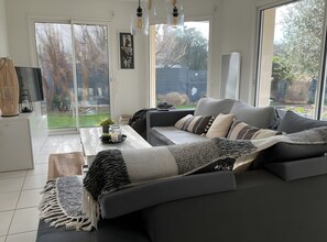 A modern living room with a grey sofa, a coffee table with decorative items, and a large window offering a view of the outdoors. - Holiday Home 'Gite Les Hauts Du Goh-Vras' with Private Pool, Private Terrace and Wi-Fi (Saint-Gildas-de-Rhuys)