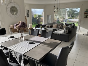 Un salon moderne avec un coin repas, un canapé gris et une vue sur le jardin grâce à de grandes fenêtres.