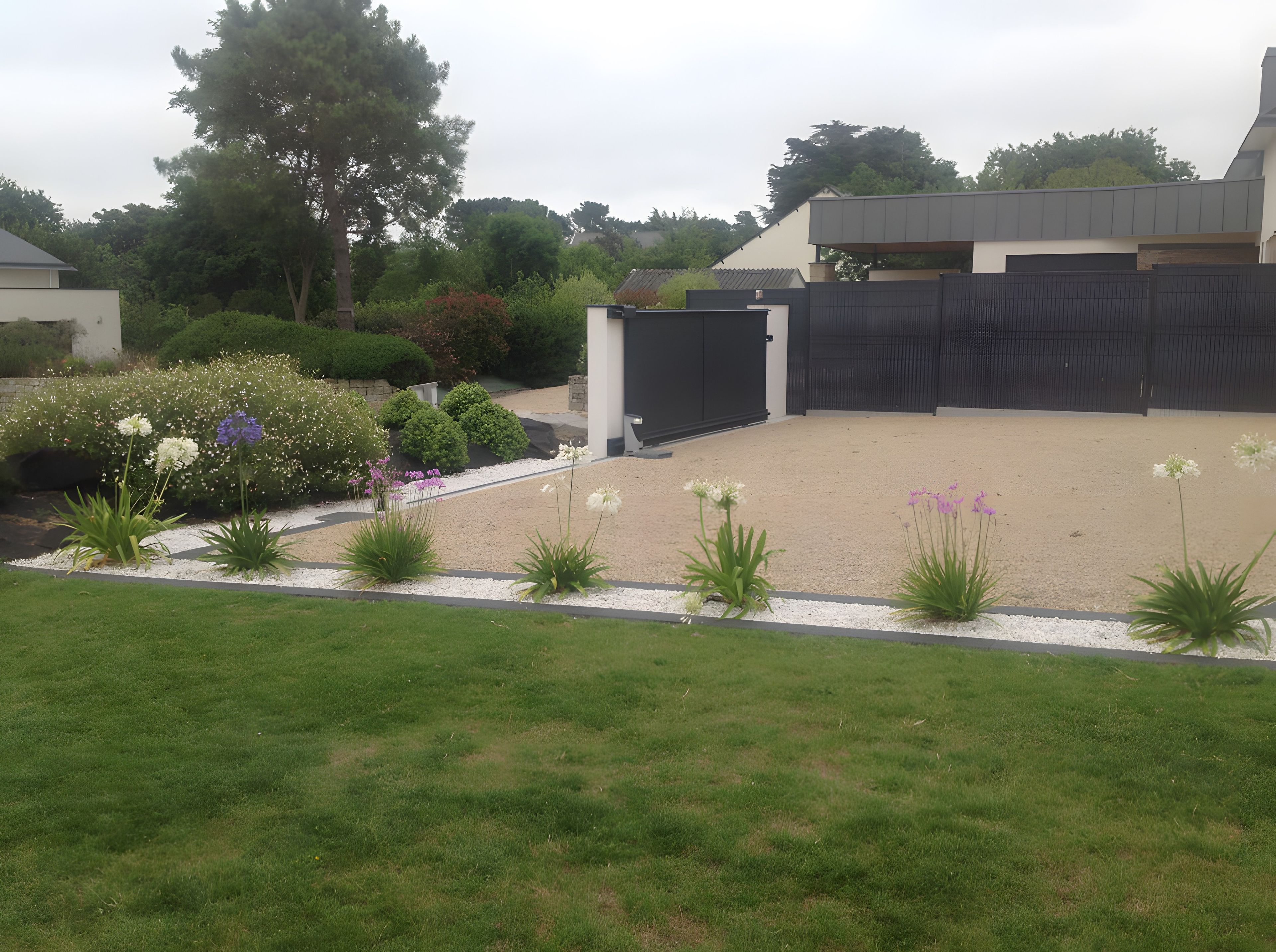 A residential garden with a gravel driveway, a variety of flowering plants, and a modern house with a dark fence.