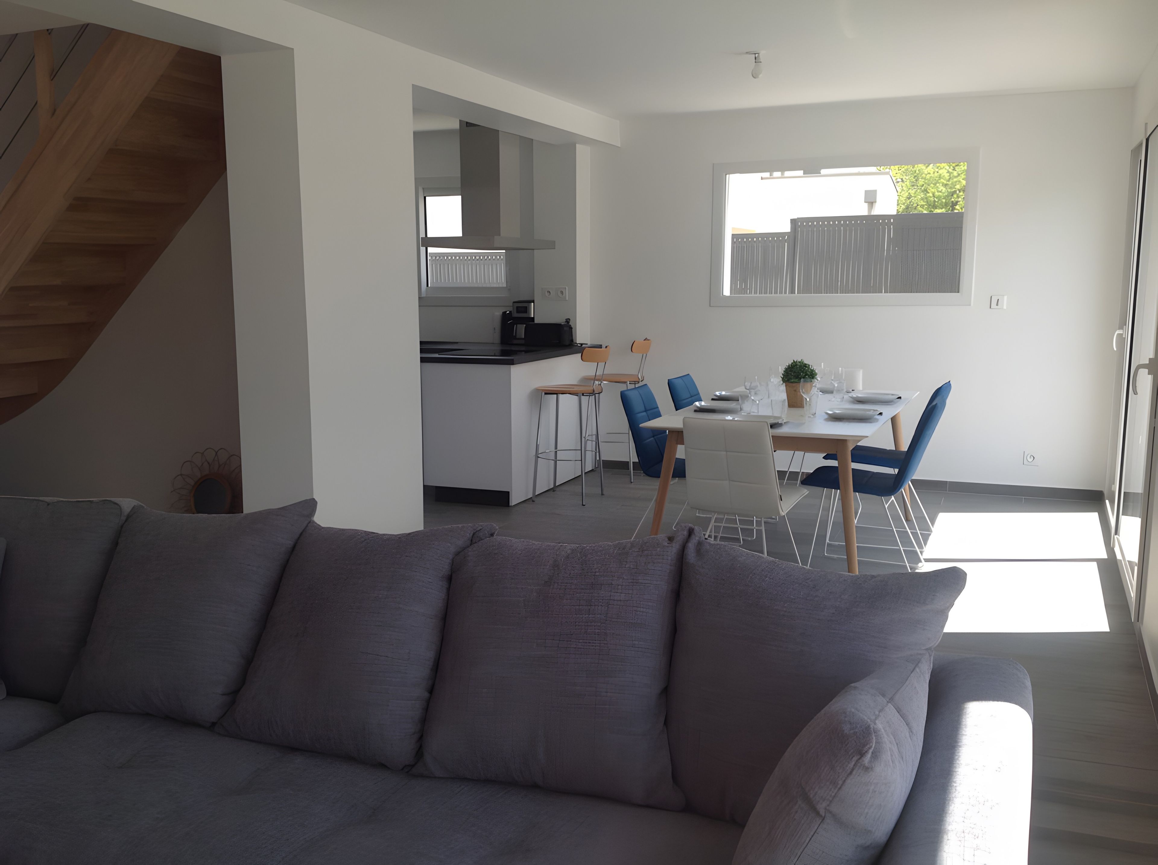 A modern living room with a grey sofa, a dining area with a table set for two, and a kitchen in the background.