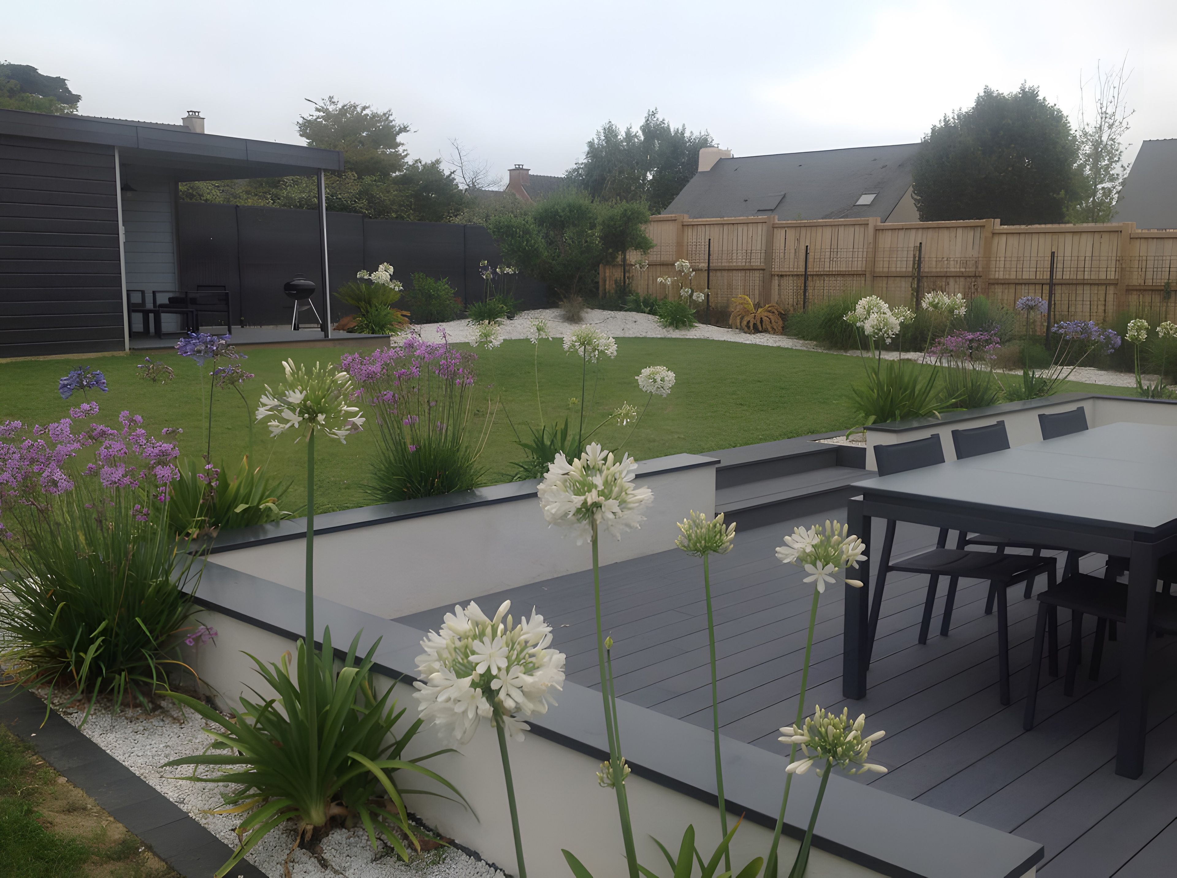 A deck with a table and chairs, surrounded by a garden with various plants and flowers.