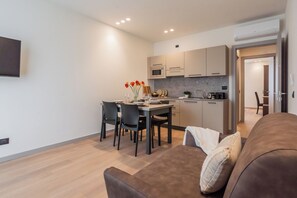 A modern living room with a brown sofa, a dining area with a table and chairs, a kitchen with a microwave and cabinets, and a TV mounted on the wall. - Close To Everything Apts (Como)