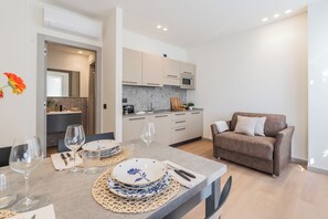 A modern kitchen with a microwave, a coffee machine, and a sink. There is a dining area with a table set for two, including plates, glasses, and cutlery. A brown sofa is positioned in the corner. - Close to Everything 1 by Rent All Como (Como)