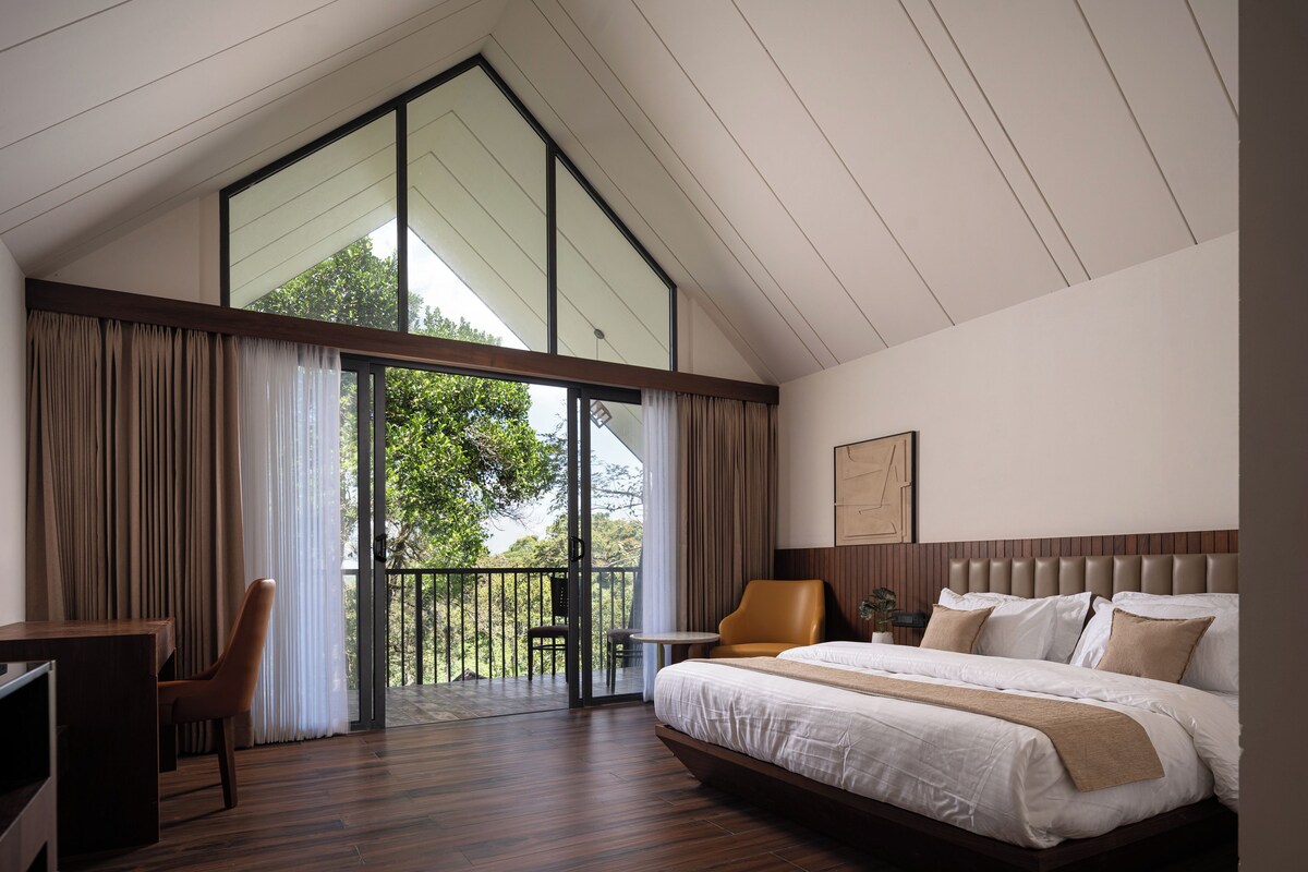 A modern bedroom with a large bed, a desk, and a chair. There is a balcony with a view of trees.