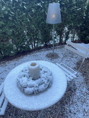 Un espace extérieur aménagé avec des sièges, recouvert de neige, comprenant une table ronde et un banc, le tout entouré d’arbres.