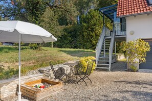 Ein Außenbereich mit Sitzgelegenheit, Tisch, Stühlen, einem Holzpflanzgefäß und einem weißen Sonnenschirm.