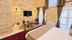 Habitación de hotel con pared de piedra, un televisor de pantalla plana sobre una pequeña mesa con sillas, una cama con cabecero de madera y un radiador.