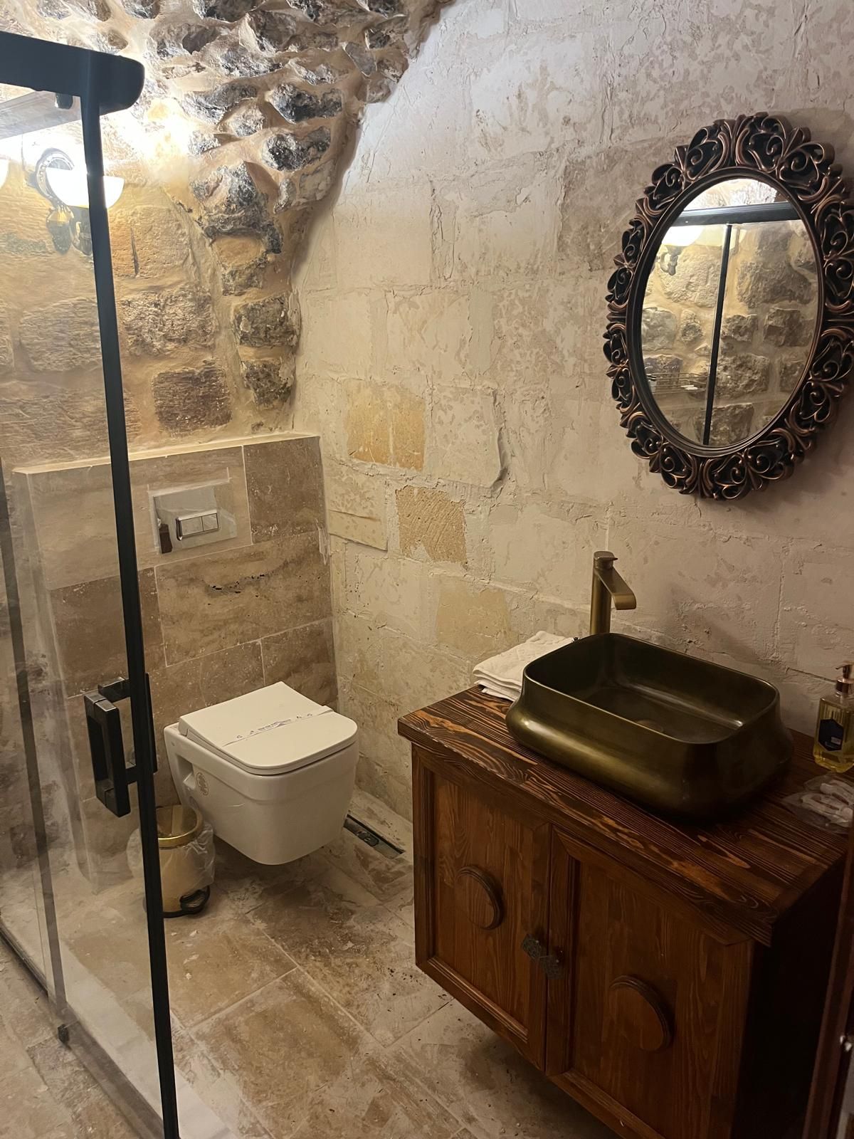 Une salle de bain avec un mur en pierre, des toilettes blanches, un meuble en bois avec un lavabo et un miroir doté d’un cadre orné.