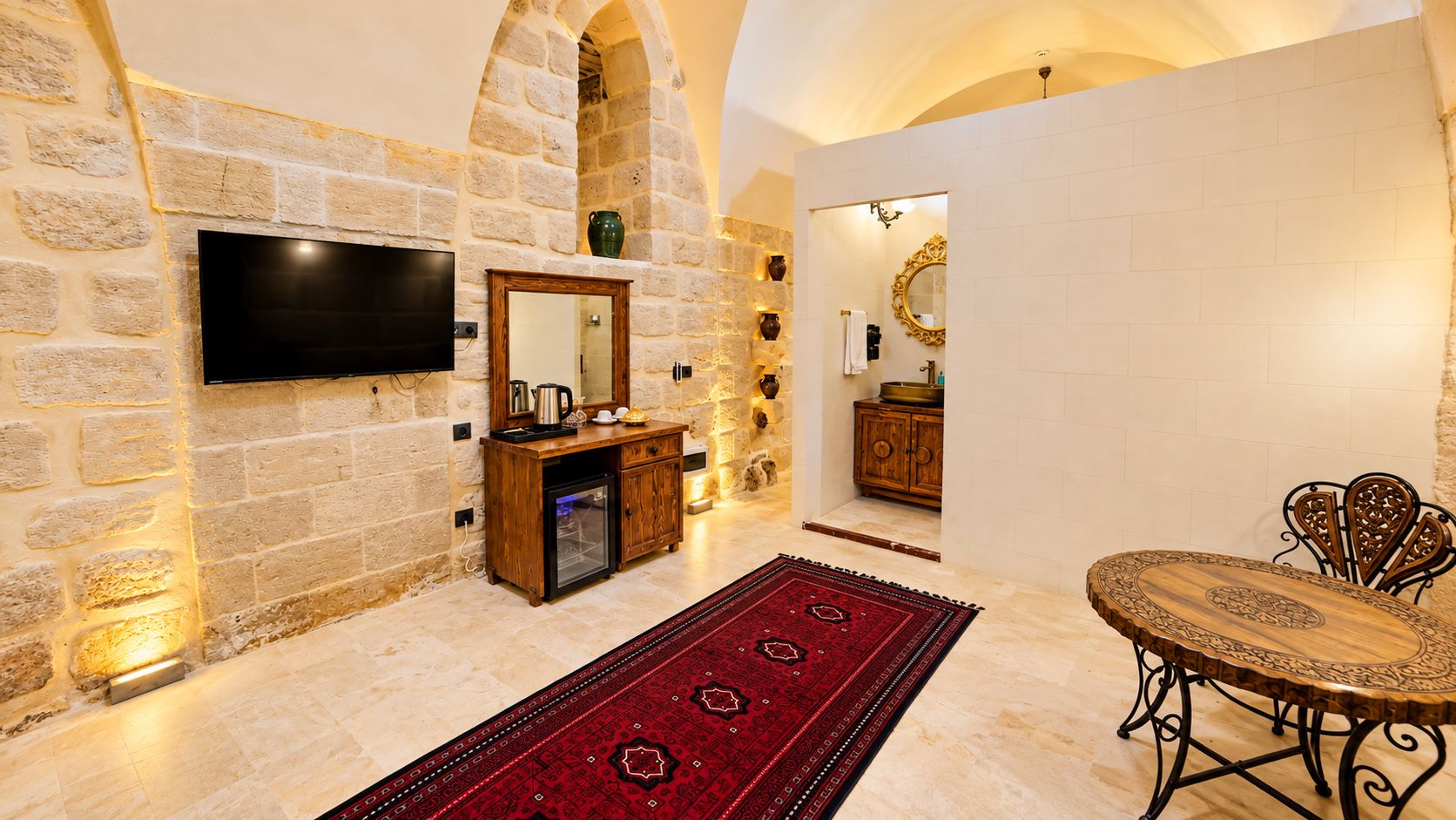 Una habitación con pared de piedra, un televisor de pantalla plana montado en ella, un mueble de madera con espejo, un tapete rojo con estampado y una mesa redonda con sillas de metal ornamentadas.