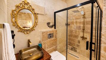 A bathroom with a gold-framed mirror, a sink with a wooden countertop, a glass-enclosed shower with a gold showerhead, and a stone wall.