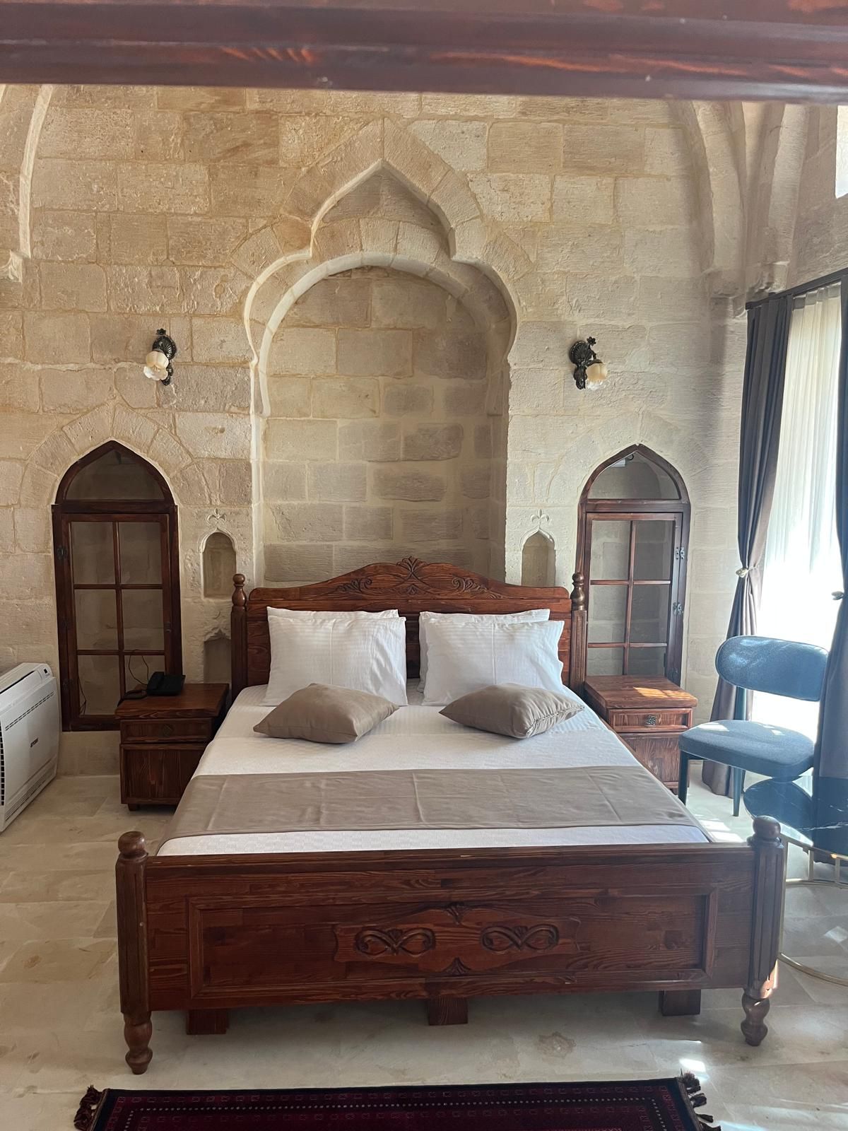 Un dormitorio con pared de piedra, una cama de madera con cabecero tallado, dos mesitas de noche con lámparas y una ventana con cortinas.