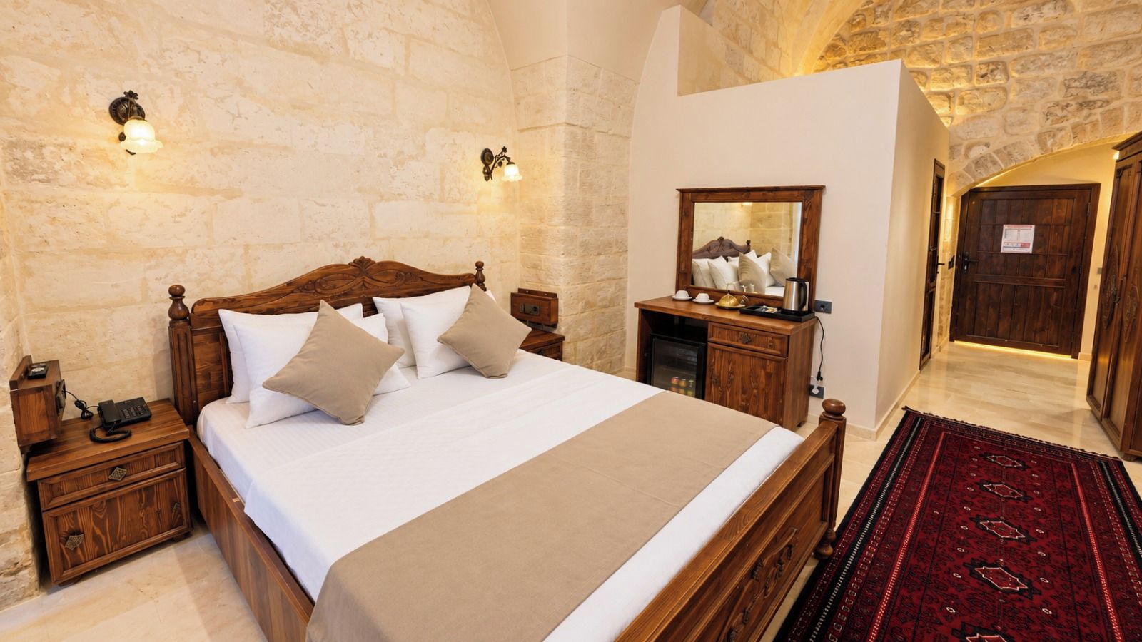 Habitación de hotel con una cama grande, mesitas de noche de madera, una mesilla, un espejo y una alfombra roja estampada.