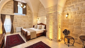 Un dormitorio con pared de piedra, cama de madera, alfombra roja y una silla de metal.