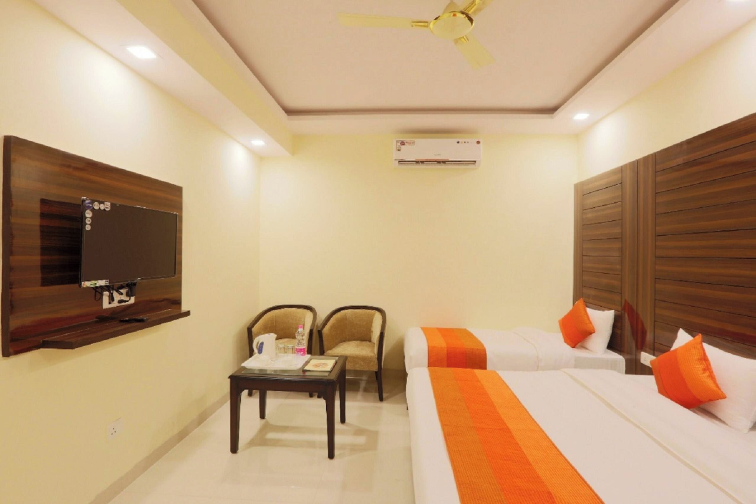 A hotel room with a bed, two chairs, a small table, a TV mounted on the wall, and an air conditioning unit.