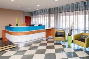Ein Hotel-Empfangsbereich mit einem Schachbrettmuster-Boden, einem blauen Empfangstresen, zwei gelben Sesseln und einem großen Fenster mit durchscheinenden Vorhängen.