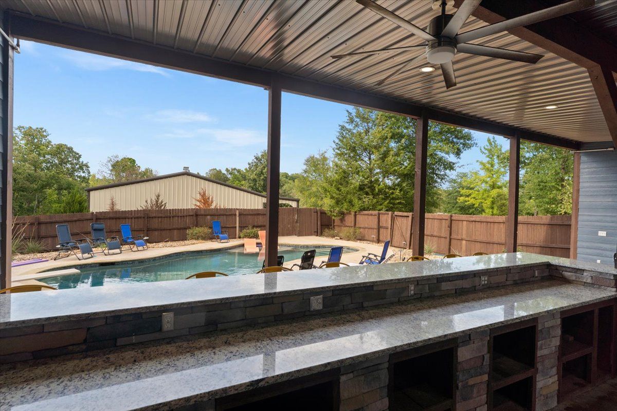 An outdoor pool area with a covered patio, stone seating, and a view of the backyard.
