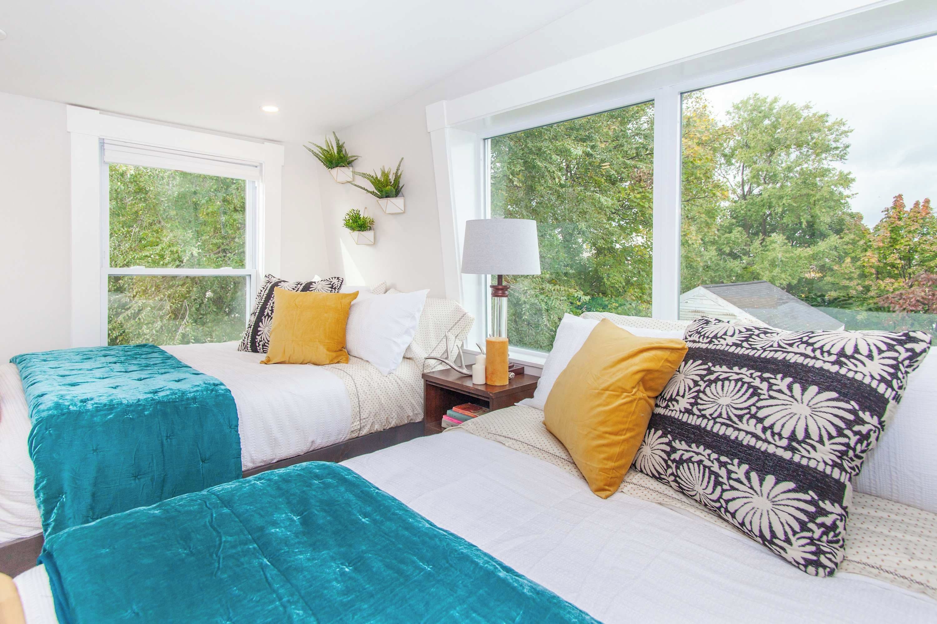 A hotel room with two beds, large windows, a bedside table with a lamp, and a view of trees.