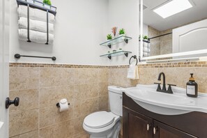 Un baño con inodoro blanco, lavamanos con dispensador de jabón, toallero con toallas dobladas y estante de vidrio con plantas.