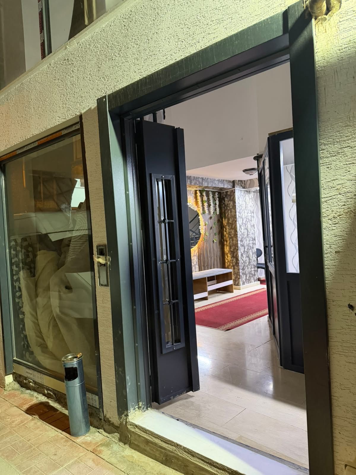 Een hotelingang met een glazen deur, een prullenbak en een doorkijkje naar een kamer met een rood tapijt en een bed.