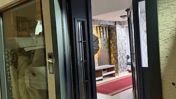 Een hotelingang met een glazen deur, een prullenbak en een doorkijkje naar een kamer met een rood tapijt en een bed.