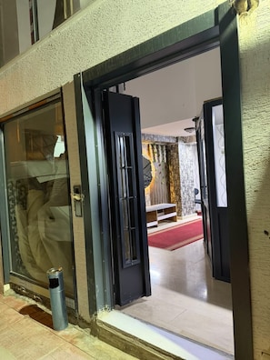 Entrada de hotel con puerta de vidrio, un contenedor de basura y vista a un cuarto con alfombra roja y una cama.