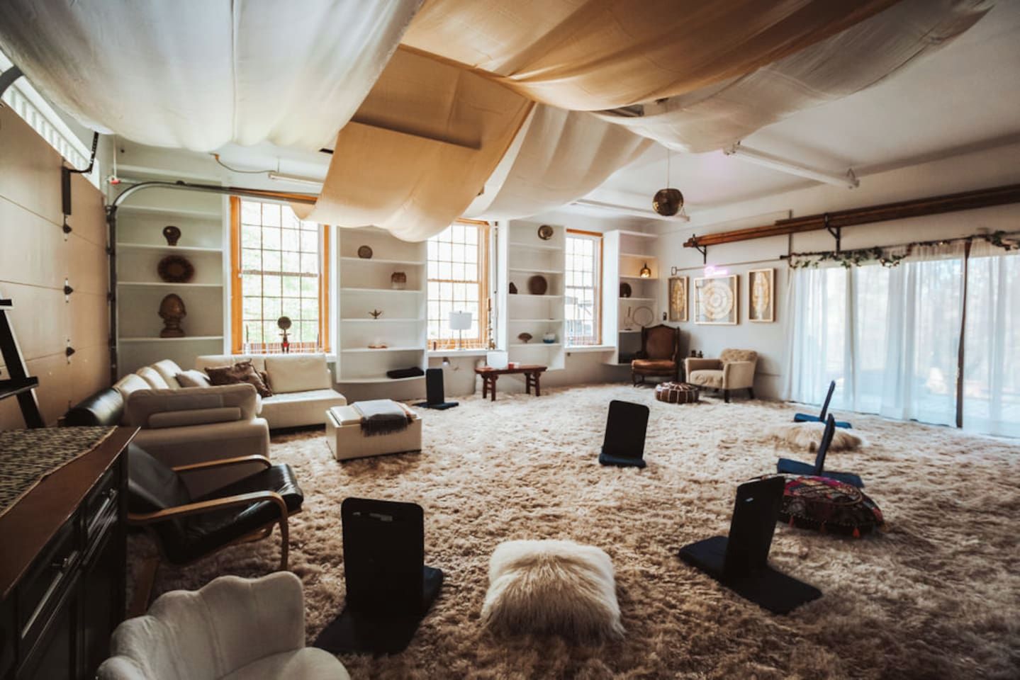 Ein geräumiger Wohnbereich mit hoher Decke, großen Fenstern und verschiedenen Sitzgelegenheiten, darunter Sofas und Sessel.